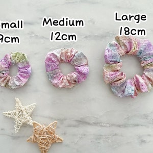 K&ouml;nnte beinhalten: Drei bunte Haargummis in verschiedenen Gr&ouml;&szlig;en: klein (9 cm), mittel (12 cm) und gro&szlig; (18 cm). Die Haargummis sind aus Stoff mit einem Meerjungfrauen-Schuppenmuster in Rosa-, Lila-, Blau- und Gr&uuml;nt&ouml;nen.