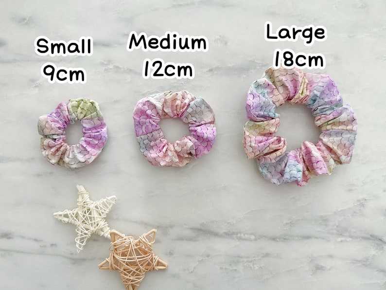 K&ouml;nnte beinhalten: Drei bunte Haargummis in verschiedenen Gr&ouml;&szlig;en: klein (9 cm), mittel (12 cm) und gro&szlig; (18 cm). Die Haargummis sind aus Stoff mit einem Meerjungfrauen-Schuppenmuster in Rosa-, Lila-, Blau- und Gr&uuml;nt&ouml;nen.