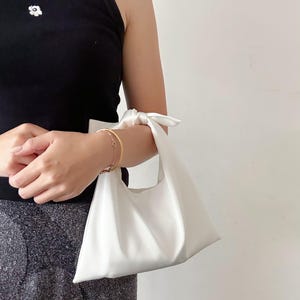May include: A small, solid white handbag featuring a smooth, matte finish and a knotted handle design. The bag has a triangular, slouchy silhouette with a single top opening, constructed from a plain, unpatterned fabric with clean, minimalist lines.