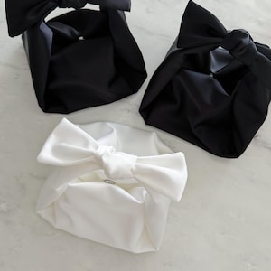 May include: Three fabric gift bags in black and white. The bags are square-shaped with a bow on top, and are made of a smooth, satin-like material. The white bag has a silver ring closure.
