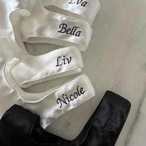 May include: A collection of satin garments featuring four white pieces embroidered with the names Eva, Bella, Liv, and Nicole in black thread, and one black piece embroidered with the name Sarah in matching black thread.