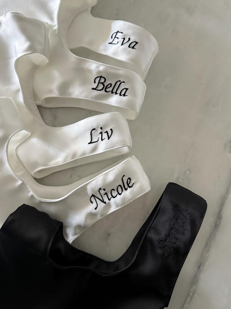 May include: A collection of satin garments featuring four white pieces embroidered with the names Eva, Bella, Liv, and Nicole in black thread, and one black piece embroidered with the name Sarah in matching black thread.