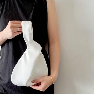 May include: A white, knotted handbag with a rounded shape. The bag is held by a person wearing a black sleeveless top. A gold bracelet and ring are also visible.