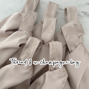 May include: A collection of beige satin champagne bags, each with a handle embroidered with initials. The bags are arranged in a close-up shot, with the text "Thread 8 on champagne bag" overlaid on the image.