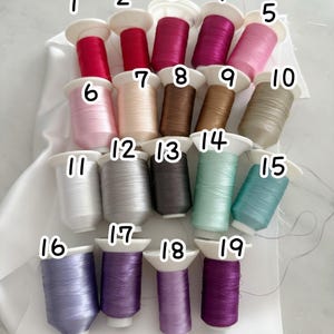 May include: A collection of 19 spools of thread in various colors, including shades of red, pink, brown, gray, green, and purple. Each spool is labeled with a number and has a white base. The threads are neatly arranged.