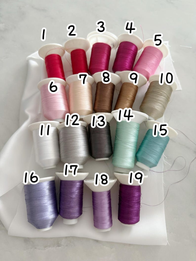 May include: A collection of 19 spools of thread in various colors, including shades of red, pink, brown, gray, green, and purple. Each spool is labeled with a number and has a white base. The threads are neatly arranged.