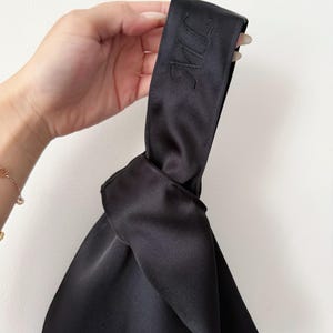 May include: Black satin handbag with a knotted design. The handle features embroidered initials. The bag has a smooth, shiny texture and is held against a white background. A gold bracelet is visible in the background.