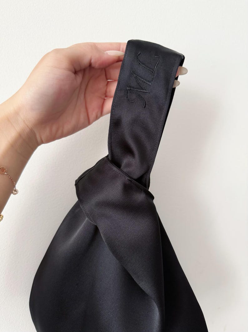May include: Black satin handbag with a knotted design. The handle features embroidered initials. The bag has a smooth, shiny texture and is held against a white background. A gold bracelet is visible in the background.