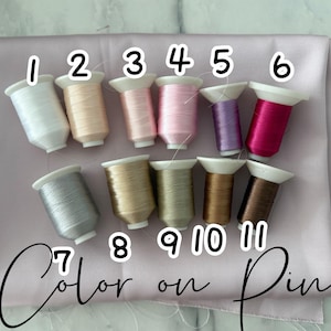 May include: An assortment of eleven spools of thread in various colors, including white, beige, pink, purple, and brown, arranged on a pink fabric. The image is labeled "Color on Pink" with numbers 1-11 above the spools.
