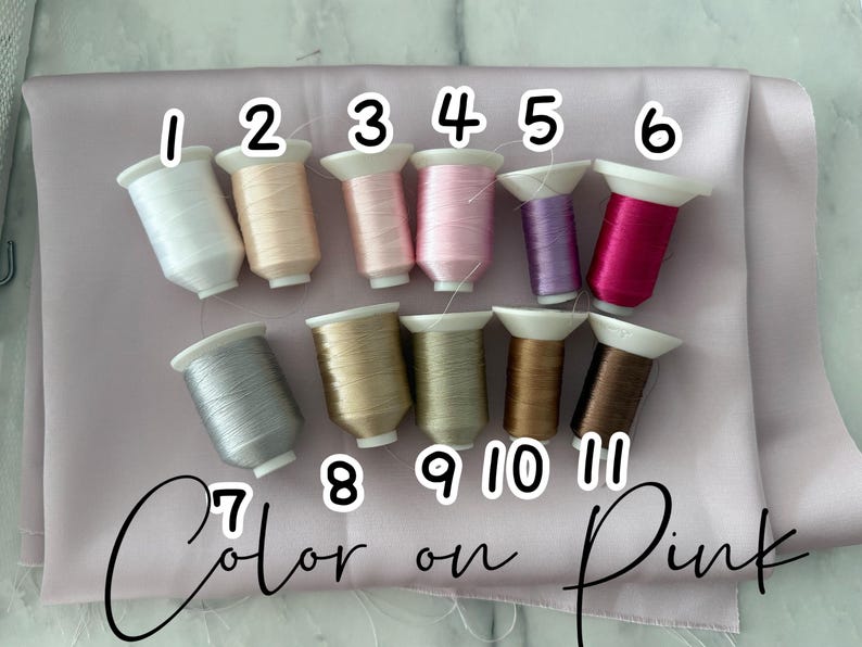 May include: An assortment of eleven spools of thread in various colors, including white, beige, pink, purple, and brown, arranged on a pink fabric. The image is labeled "Color on Pink" with numbers 1-11 above the spools.