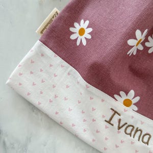 May include: A pink drawstring bag with a white daisy print and a white bottom with a pink heart print. The bag has the name "Ivana" embroidered on the bottom.