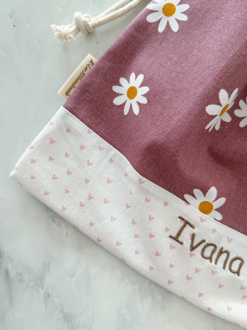 May include: A pink drawstring bag with a white daisy print and a white bottom with a pink heart print. The bag has the name "Ivana" embroidered on the bottom.