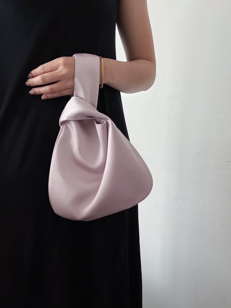May include: A light purple satin handbag with a knotted handle. The bag is held by a person wearing a black dress. The bag's shape is rounded and soft, with a smooth, shiny texture. The handle is a continuation of the bag's material.