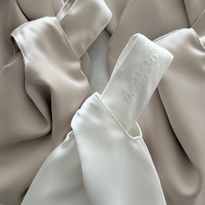 May include: A white satin knot bag featuring the embroidered date 6.13.26 on the handle, surrounded by several beige satin knot bags. The bags have a smooth, lustrous finish and a distinctive knotted handle design with soft, draped fabric.