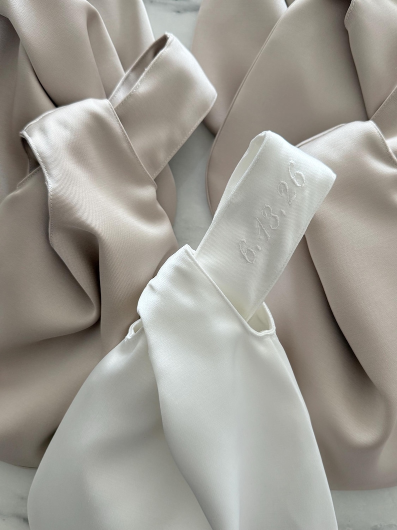 May include: A white satin knot bag featuring the embroidered date 6.13.26 on the handle, surrounded by several beige satin knot bags. The bags have a smooth, lustrous finish and a distinctive knotted handle design with soft, draped fabric.