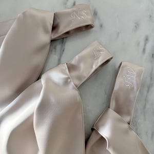 May include: Three champagne-colored satin headbands with embroidered initials. The headbands are knotted and have a soft, lustrous sheen. The initials "NS", "LS", and "MW" are embroidered in white thread.