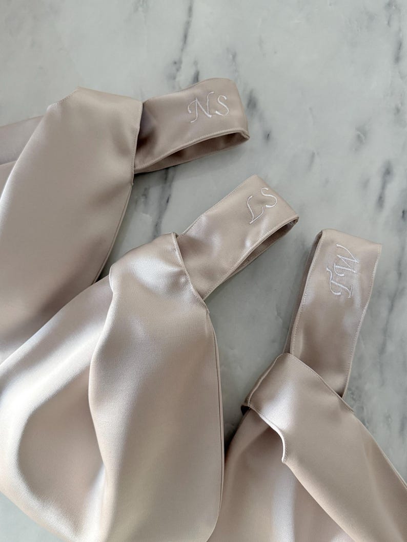 May include: Three champagne-colored satin headbands with embroidered initials. The headbands are knotted and have a soft, lustrous sheen. The initials "NS", "LS", and "MW" are embroidered in white thread.