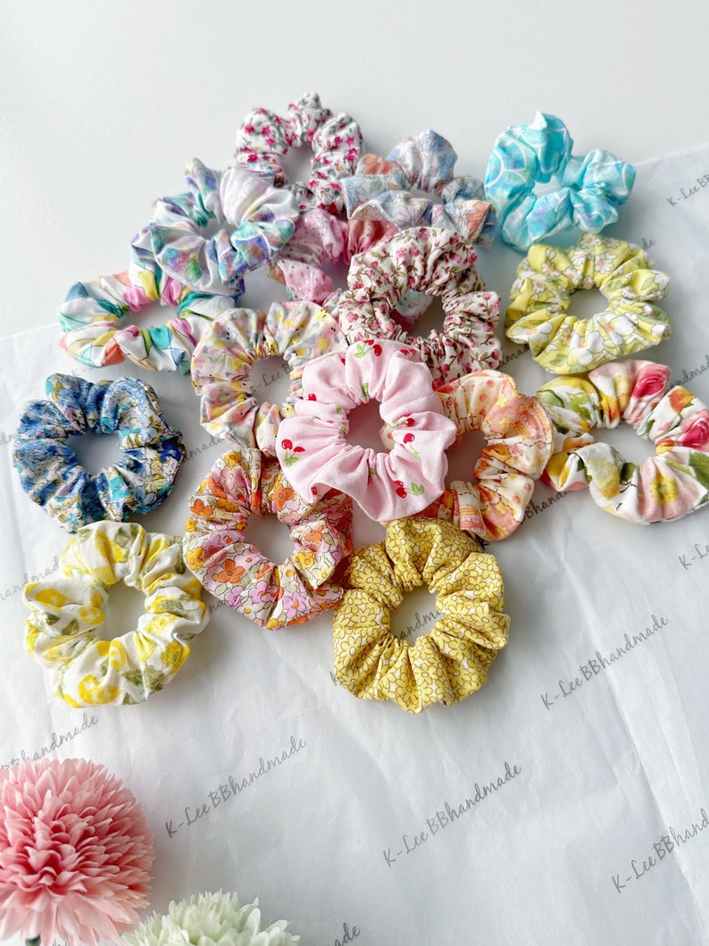 K&ouml;nnte beinhalten: Eine Sammlung von 15 bunten Stoffscrunchies mit Blumen- und anderen Mustern. Die Scrunchies sind auf einer wei&szlig;en Oberfl&auml;che zu einem Stapel angeordnet. Die Scrunchies sind handgefertigt und haben eine Vielzahl von Farben und Mustern.