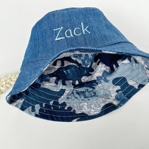May include: A blue denim bucket hat with a dinosaur print lining. The hat is embroidered with the name "Zack".