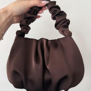 May include: A dark brown satin handbag featuring a soft, gathered, and pleated texture throughout the body. The bag includes a single, matching dark brown ruched handle that creates a ruffled appearance, providing a cohesive and uniform look.