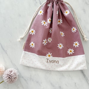 May include: A pink drawstring bag with a white daisy pattern and a white bottom with pink hearts. The bag is personalized with the name "Ivana" in brown embroidery.
