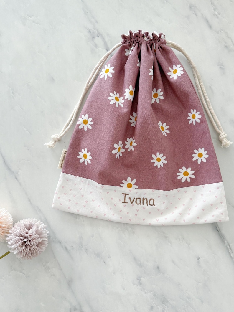 May include: A pink drawstring bag with a white daisy pattern and a white bottom with pink hearts. The bag is personalized with the name "Ivana" in brown embroidery.