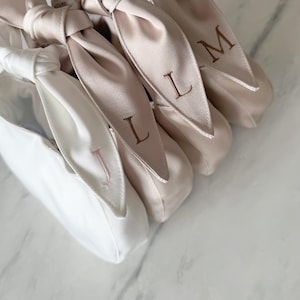 May include: A collection of three satin pouches featuring knotted top handles. One pouch is white with a pink embroidered letter J, while the other two are champagne-coloured with brown embroidered letters L and M on the knotted handles.