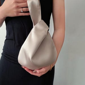 May include: A beige satin handbag with a knotted handle. The bag has a rounded shape and is held by a person wearing a black dress. The bag's color is a neutral tone, suitable for various outfits.