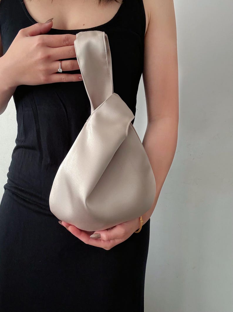 May include: A beige satin handbag with a knotted handle. The bag has a rounded shape and is held by a person wearing a black dress. The bag's color is a neutral tone, suitable for various outfits.