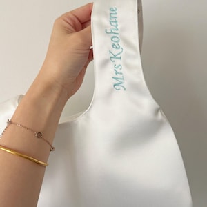 May include: A white satin handbag featuring a single integrated handle. The handle is embroidered with the text Mrs Keohane in a light blue script font. The bag has a smooth, lustrous finish and a simple, minimalist structural design.