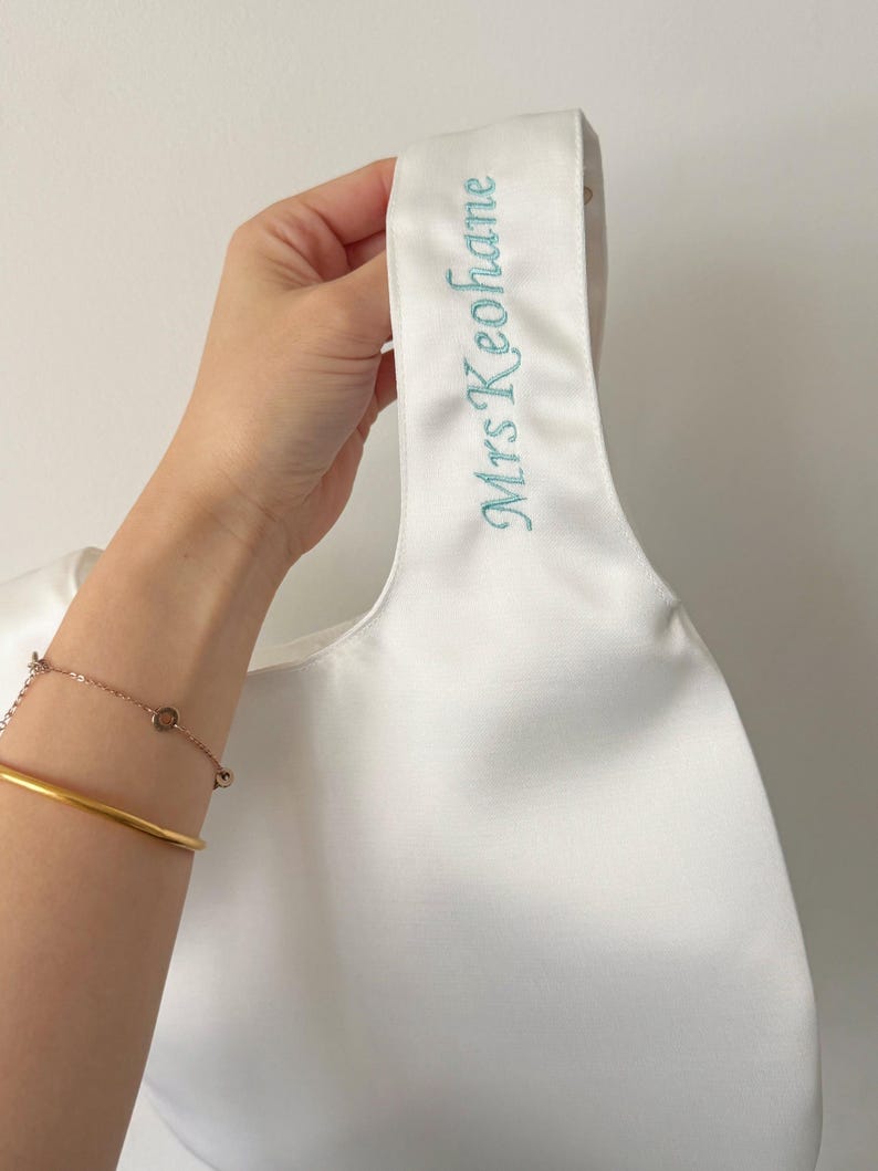 May include: A white satin handbag featuring a single integrated handle. The handle is embroidered with the text Mrs Keohane in a light blue script font. The bag has a smooth, lustrous finish and a simple, minimalist structural design.