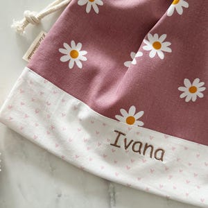 May include: A pink drawstring bag with a white daisy print and a white border with pink hearts. The bag is personalized with the name "Ivana".