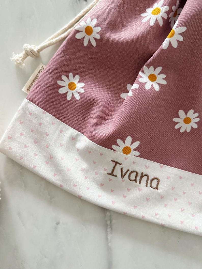 May include: A pink drawstring bag with a white daisy print and a white border with pink hearts. The bag is personalized with the name "Ivana".