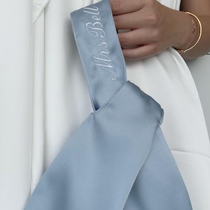 Op de afbeelding: Een lichtblauwe satijnen sjerp met de geborduurde tekst "Mrs. Bell" in witte letters. De sjerp wordt vastgehouden door een persoon die een witte jurk draagt. Een gouden armband is zichtbaar om de pols van de persoon.