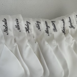 May include: A row of white satin pouches, each featuring a name embroidered in black cursive script. The names visible are Katie, Natalie, Alexis, Haley, Abby, Morgan, Bailey, Hannah, Jordan, and Olivia, all displayed in a uniform font.