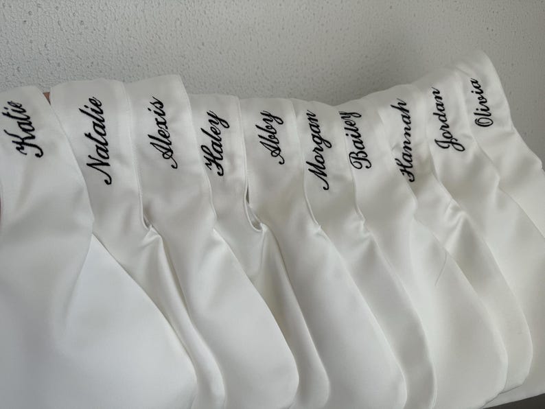 May include: A row of white satin pouches, each featuring a name embroidered in black cursive script. The names visible are Katie, Natalie, Alexis, Haley, Abby, Morgan, Bailey, Hannah, Jordan, and Olivia, all displayed in a uniform font.