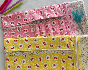 May include: Two fabric organizers for crochet hooks, one in pink and one in yellow, both featuring a repeating strawberry pattern. Each organizer includes multiple slots for hooks, a zippered side pocket, and a floral-patterned interior lining.