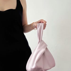 May include: A light pink satin handbag with a knotted handle. The bag has a triangular shape and is held by a person wearing a black dress. The bag's smooth texture and soft color create a minimalist aesthetic.