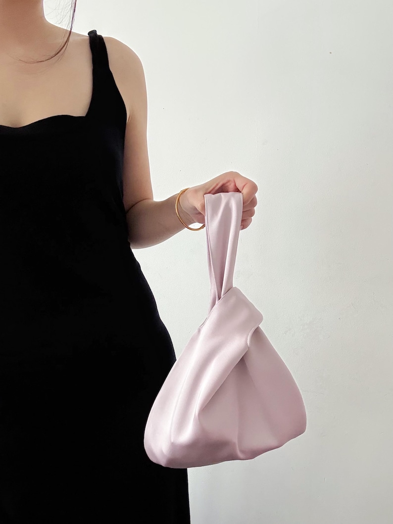 May include: A light pink satin handbag with a knotted handle. The bag has a triangular shape and is held by a person wearing a black dress. The bag's smooth texture and soft color create a minimalist aesthetic.