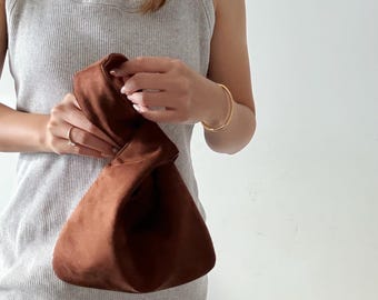Könnte beinhalten: Eine braune Wildleder-Clutch-Tasche mit einem einzigartigen Knoten-Design. Die Tasche wird von einer Person gehalten, die ein graues geripptes Tanktop und ein goldenes Armband trägt. Die Farbe der Tasche ist ein sattes, warmes Braun und wirkt weich.
