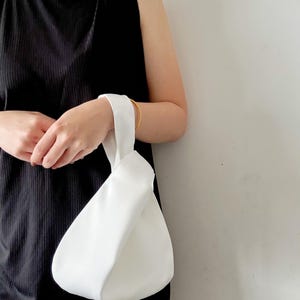 May include: A solid white knot bag featuring a single handle loop design. The bag has a smooth, matte texture and a rounded, teardrop-shaped silhouette. The handle is formed by pulling one side of the bag through the other, creating a secure closure.