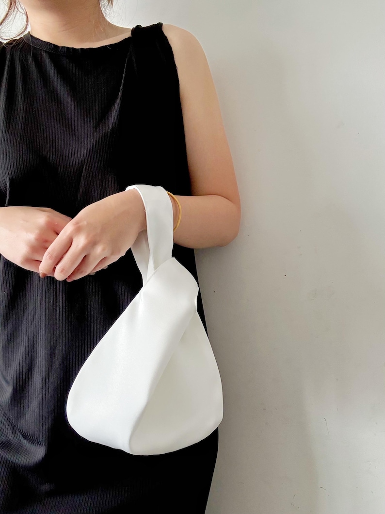 May include: A solid white knot bag featuring a single handle loop design. The bag has a smooth, matte texture and a rounded, teardrop-shaped silhouette. The handle is formed by pulling one side of the bag through the other, creating a secure closure.