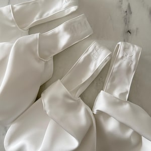 May include: Four white satin bags with embroidered initials on the handles. The bags are gathered at the base and have a smooth, shiny texture. The initials "TC", "AM", and "CS" are visible.
