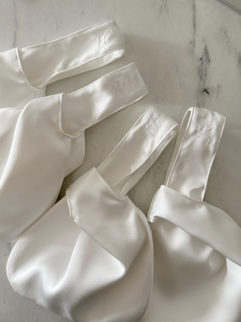 May include: Four white satin bags with embroidered initials on the handles. The bags are gathered at the base and have a smooth, shiny texture. The initials "TC", "AM", and "CS" are visible.