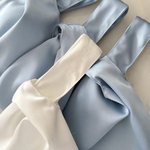 May include: Three knot bags made of smooth satin fabric. Two bags are light blue and one bag is white. The white bag features the embroidered text 18.6.25 on the handle. Each bag has a single handle formed by a knot design.