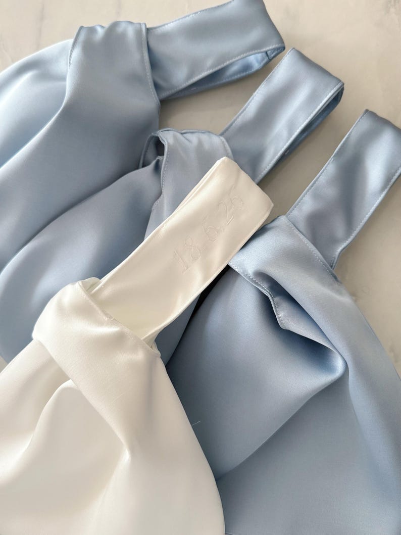 May include: Three knot bags made of smooth satin fabric. Two bags are light blue and one bag is white. The white bag features the embroidered text 18.6.25 on the handle. Each bag has a single handle formed by a knot design.