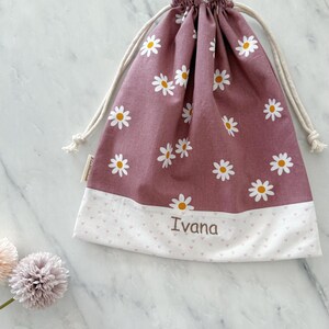 May include: A pink drawstring bag with a white daisy print and a white bottom with pink hearts. The bag is personalized with the name "Ivana" in brown embroidery.