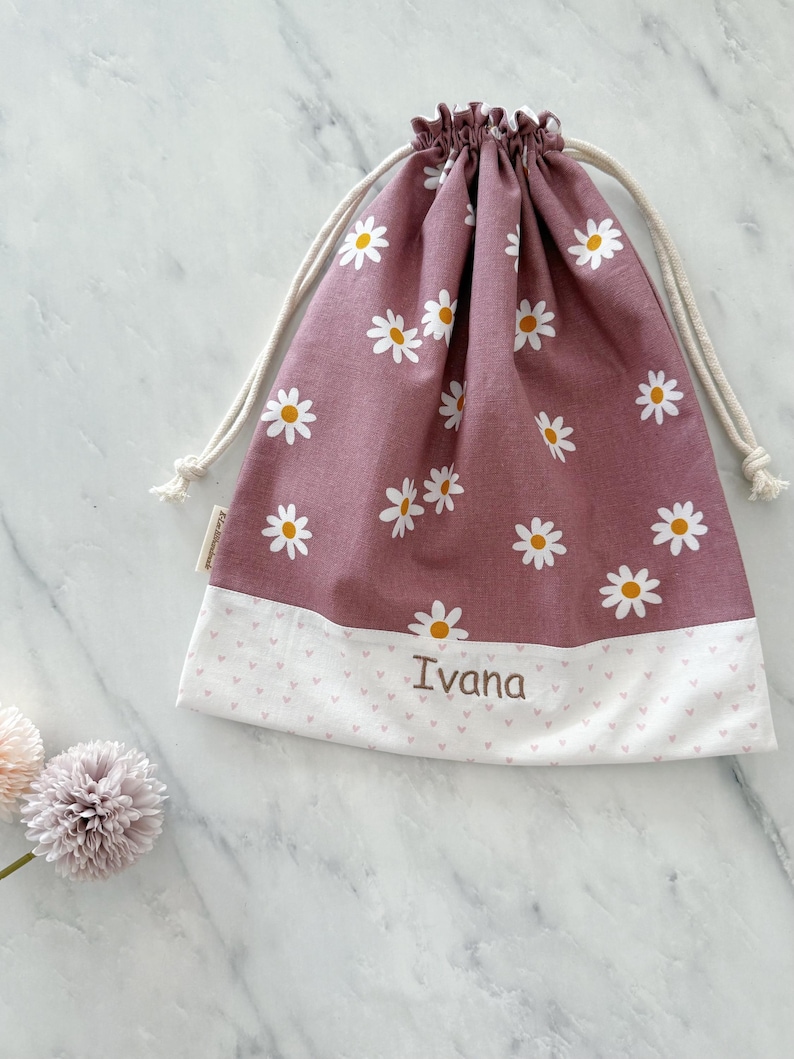 May include: A pink drawstring bag with a white daisy print and a white bottom with pink hearts. The bag is personalized with the name "Ivana" in brown embroidery.