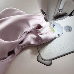 May include: A close-up shot of a sewing machine in operation, stitching together a piece of pale pink fabric. The fabric is gathered and being fed through the machine, with the needle and presser foot clearly visible. The machine is on a light-colored surface.