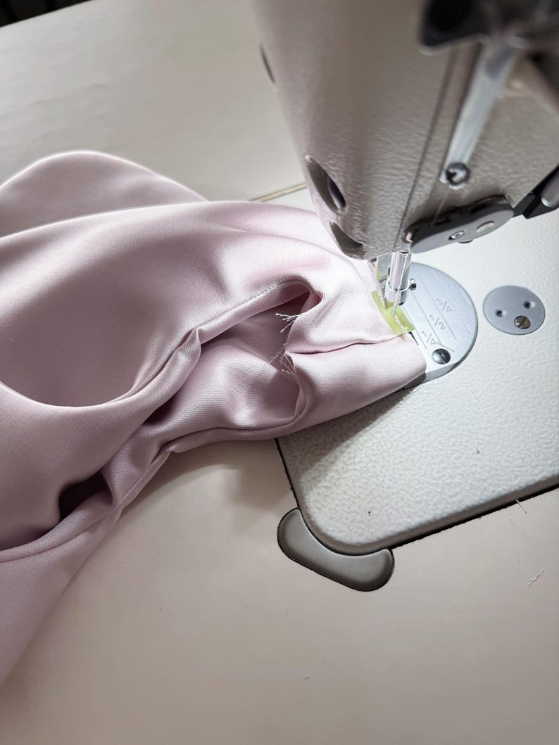May include: A close-up shot of a sewing machine in operation, stitching together a piece of pale pink fabric. The fabric is gathered and being fed through the machine, with the needle and presser foot clearly visible. The machine is on a light-colored surface.
